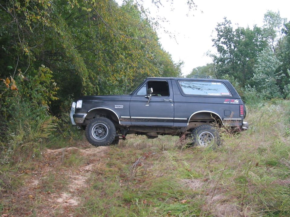 Dark blue Ford Bronco driving on a dirt path with trees and tall grass around it