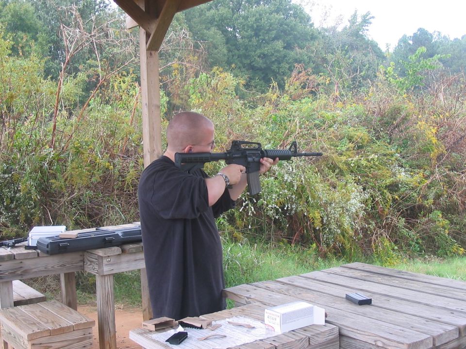 Man aims rifle at outdoor shooting range with trees and brush in background