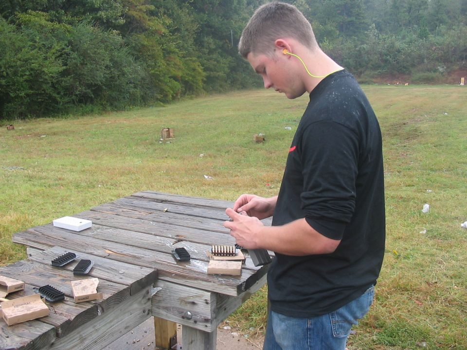 guy loading bullets into a magazine at a wooden table on a grassy range