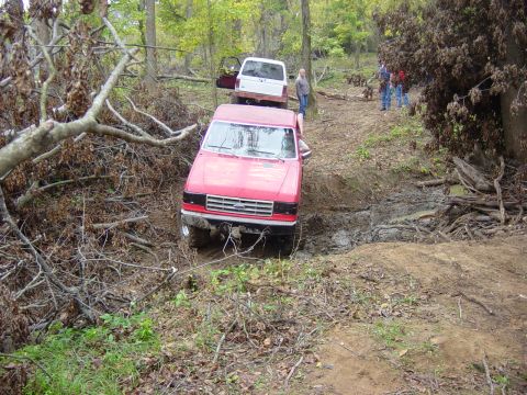 1983 Ford Bronco - A red pickup truck navigates a muddy off-road trail with a white vehicle following behind.