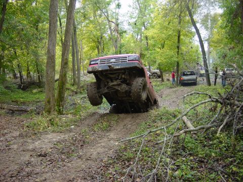 1983 Ford Bronco - Red SUV navigating a challenging off-road trail in a wooded area with other vehicles present.