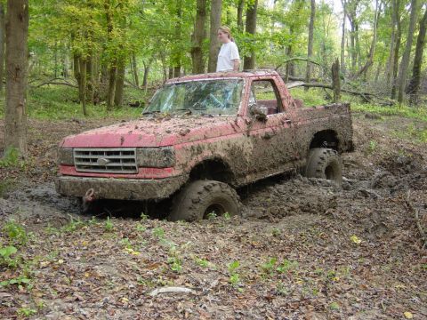 1983 Ford Bronco - A red pickup truck is stuck in thick mud while off-roading in a wooded area.