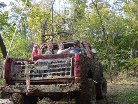 1983 Ford Bronco - A group of people ride in the back of a heavily mud-covered red pickup truck through a wooded area.
