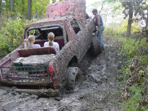 1983 Ford Bronco - A heavily mud-caked vehicle is stuck in a wooded area with two passengers inside and another person attempting to push it free.