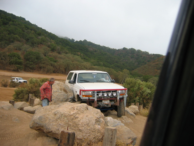 White and red lifted truck with round driving lights crawling boulders as a man watches in fog