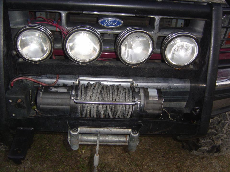 Front view of a dark red Ford Bronco with a black brushguard, four auxiliary lights, and a winch. The Ford logo is visible on the grille