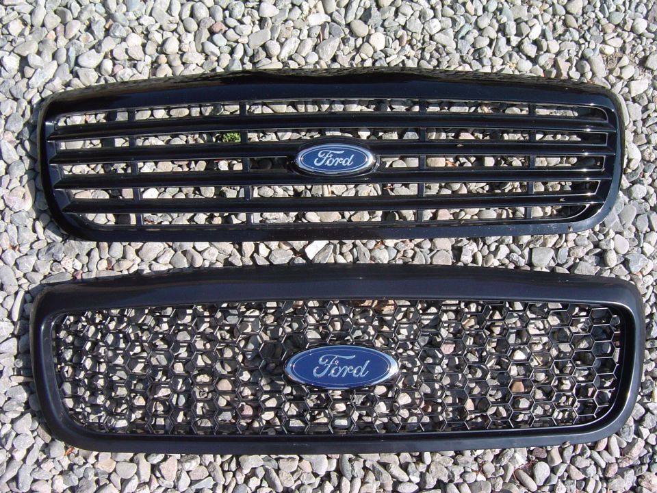 Two black Ford grilles with blue oval emblems lying on gravel, one slatted and one honeycomb mesh