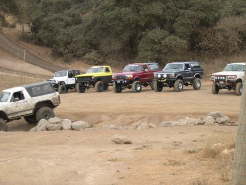 1990 Ford Bronco - A group of modified off-road vehicles are parked on a dusty trail with trees in the background.