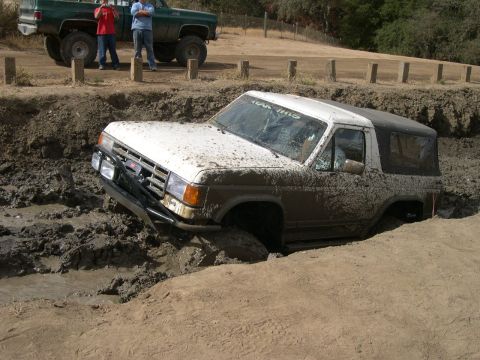 1990 Ford Bronco - A white and brown SUV is stuck in a mud pit, while a green pickup truck with two people stands by.