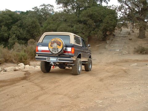 1990 Ford Bronco - 1990 Ford Bronco navigating an off-road trail with trees and hills in the background.
