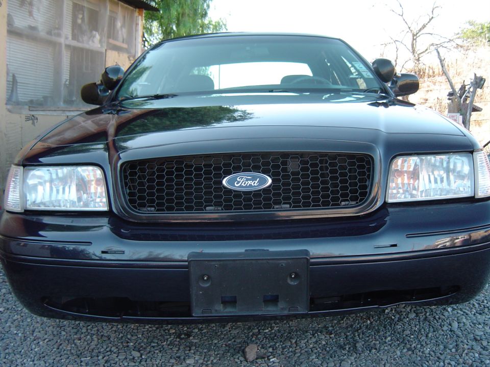 Front view of a dark Ford Crown Victoria with black grille and empty license plate bracket on gravel