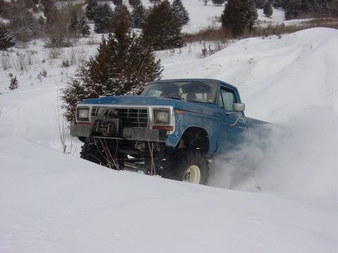 1983 Ford Bronco - A lifted blue pickup truck navigates a snowy terrain, its tires churning up a cloud of snow.