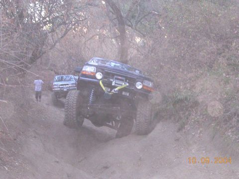 1994 Ford Bronco - Two SUVs navigate a dusty, wooded trail with steep inclines and loose dirt.