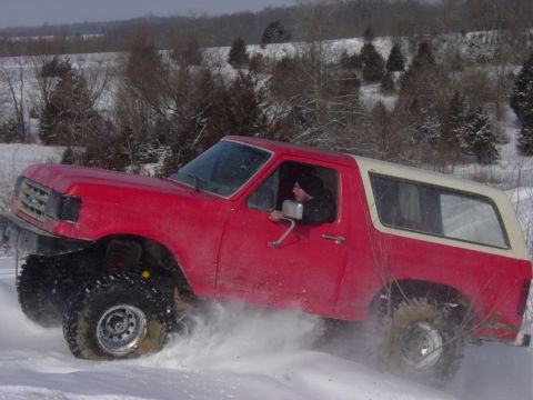 1983 Ford Bronco - A red SUV with a white top navigates a snowy terrain, sending snow flying.