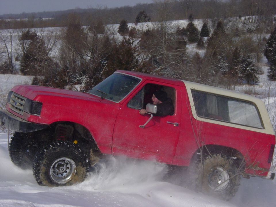 Lifted red Ford Bronco with white top blasting through deep snow, driver leaning out window