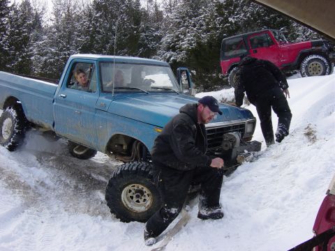 1983 Ford Bronco - A blue pickup truck is stuck in a snowy ditch, with a red Jeep visible behind it.
