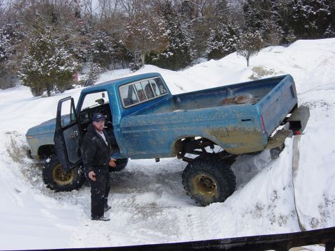 1983 Ford Bronco - A man stands beside a heavily modified blue pickup truck stuck in the snow.