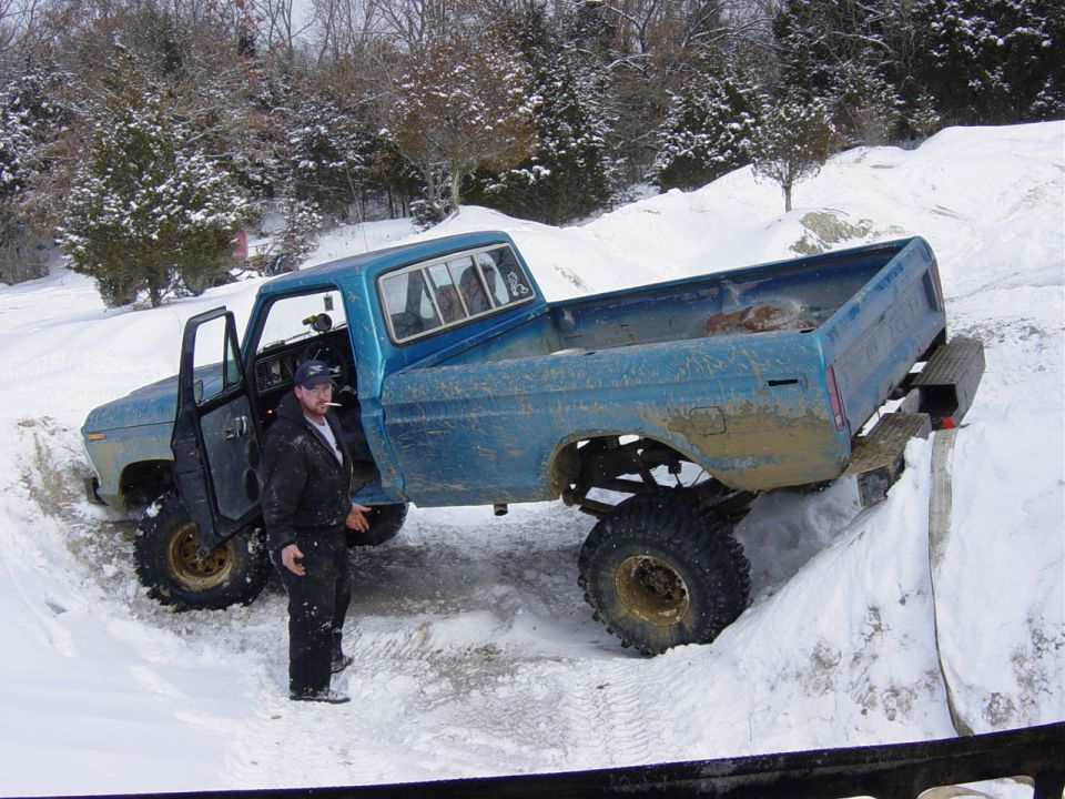 Muddy lifted blue Ford pickup stuck in a snow trench with a man standing at the open door