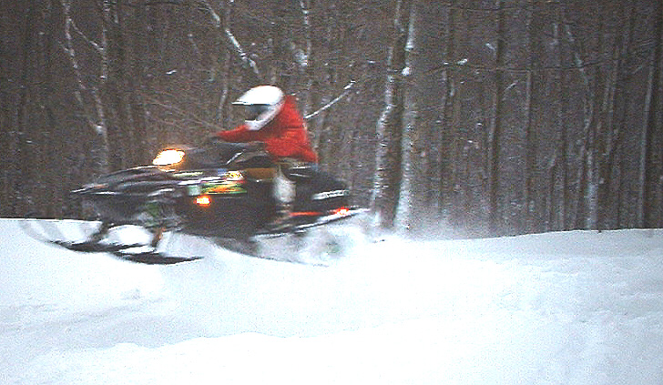 Rider in red jacket jumps a dark snowmobile over snow with bare trees behind