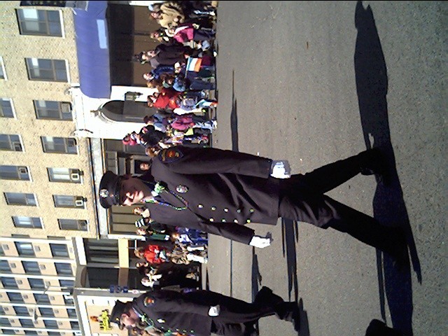 Uniformed parade marchers wearing beads walk past spectators along a street; image rotated sideways