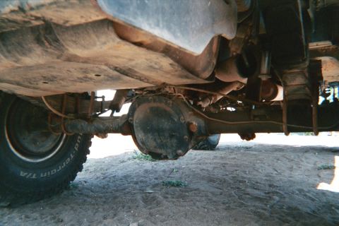 1990 Ford Bronco - Low underbody view of a 4x4 showing a dusty front differential, axle, driveline and a BFGoodrich mud-terrain tire on sandy ground.