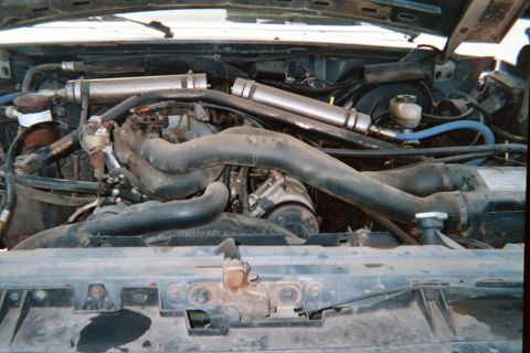 1990 Ford Bronco - engine bay fitted with twin polished metal tubes routed over a V-engine, with hoses, brackets and a bare front radiator support visible.