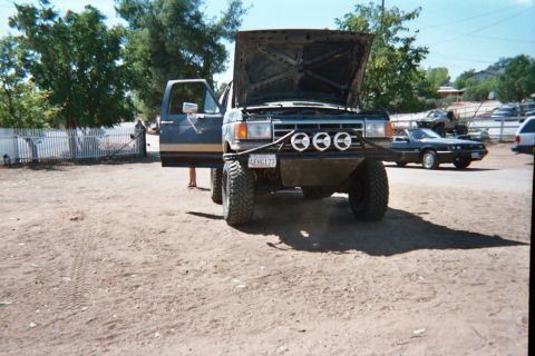1990 Ford Bronco - Front view of a lifted dark Ford SUV with its hood raised and a four-light bumper bar, parked in a fenced dirt yard with other cars.
