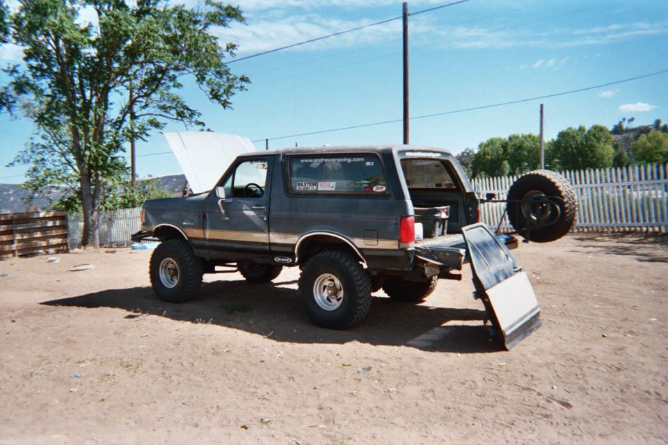 Lifted dark Ford Bronco with gold stripe and rear spare parked in a dirt yard