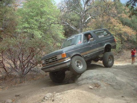 1990 Ford Bronco - lifted dark Ford Bronco climbs over a large rock on a brushy wooded trail while a person in red looks on from the side.