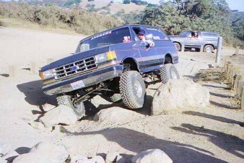 1990 Ford Bronco - lifted Ford Bronco flexes its suspension over boulders on a dirt trail, with a second Bronco waiting in the background.