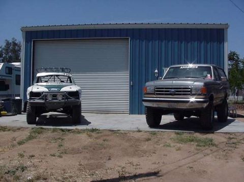 1990 Ford Bronco - white tube-front prerunner truck and a lifted Ford Bronco are parked side by side in front of a blue corrugated metal shop building.