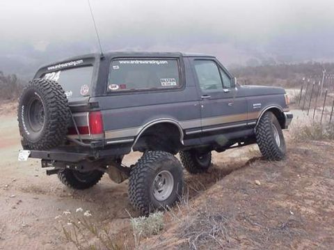 1990 Ford Bronco - Rear-quarter view of a lifted dark Ford Bronco with a tailgate spare and racing decals, parked on a foggy open dirt trail.