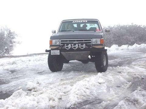 1990 Ford Bronco - Front view of a dark Ford Bronco with a four-light bumper bar parked on a snow-covered dirt road under an overcast sky.