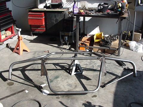 1990 Ford Bronco - bare welded tubular metal frame or roll structure sits on jack stands on a shop floor, with a workbench and tool chest behind.