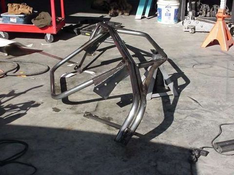 1990 Ford Bronco - fabricated bare-steel tubular assembly sits on a concrete shop floor, with jack stands, a red tool cart, and a dog in the background.