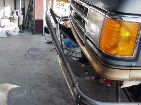 1990 Ford Bronco - black tubular bumper is fitted beneath the grille and amber corner lamp of a gray vehicle inside a workshop.