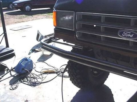 1990 Ford Bronco - black tubular bumper mounts to a lifted truck bearing a Ford grille emblem, with a welding helmet and leads scattered on the driveway.