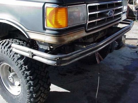 1990 Ford Bronco - bare-steel tubular bumper is bolted beneath the grille and amber lamp of a gray-and-tan lifted Ford truck on mud tires.
