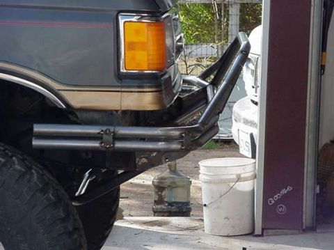1990 Ford Bronco - Side angle of a bare-steel tubular bumper on a lifted Ford truck parked by a garage door, with plastic buckets and a water jug on the groun…