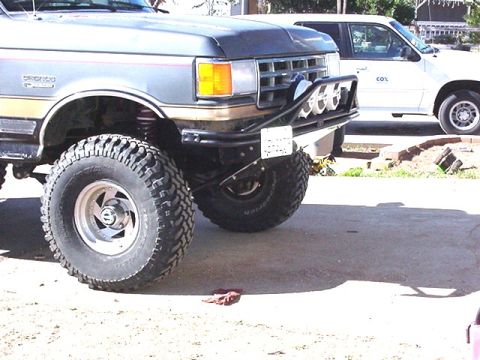 1990 Ford Bronco - Three-quarter front view of a lifted gray Ford truck on tall mud tires, fitted with a tubular bumper and round driving lights.