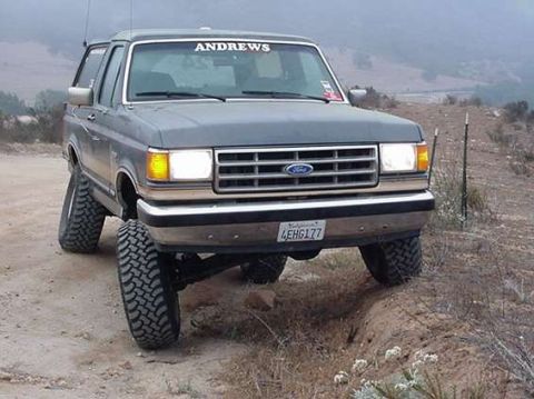 1990 Ford Bronco - gray lifted Ford Bronco on mud tires sits on a dirt trail before rolling hills, with a Ford grille emblem and an ANDREWS windshield banner.