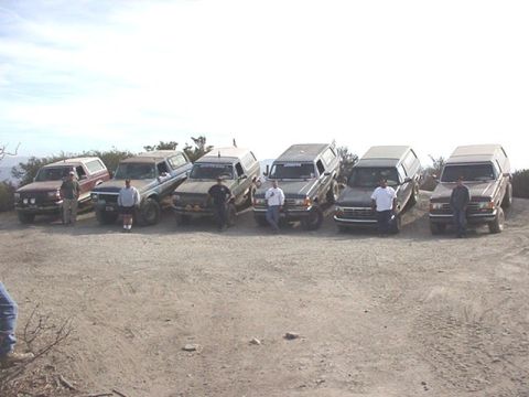 1990 Ford Bronco - Six SUVs and trucks are lined up with people standing beside them on a gravel hilltop overlook under an overcast sky.