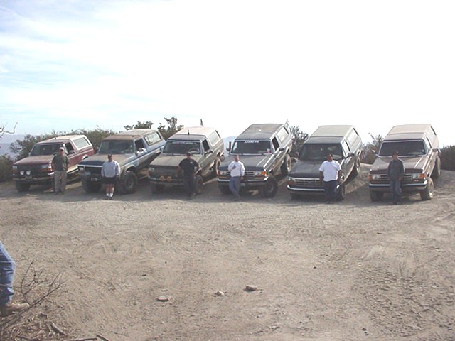 Row of six SUVs and trucks with people standing beside them on a gravel hilltop overlook