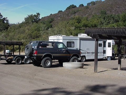 1990 Ford Bronco - lifted black-and-tan Ford Bronco with a spare tire on the tailgate is parked beside a white Itasca motorhome at a dirt campsite.