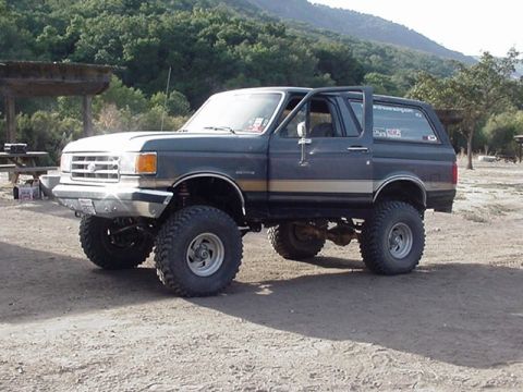 1990 Ford Bronco - lifted gray-and-tan Ford Bronco on mud tires with a tubular front bumper is parked on a dirt lot backed by green hills.