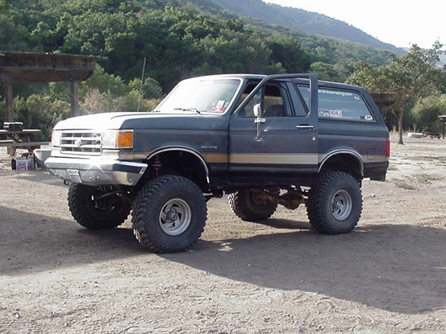 Lifted gray-and-tan Ford Bronco with tube bumper parked on dirt with green hills behind