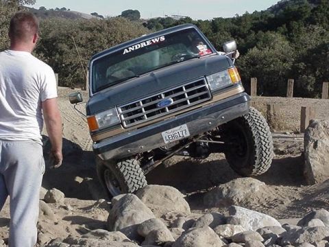 1990 Ford Bronco - lifted Ford truck with an ANDREWS windshield banner climbs a rock ledge on a dirt trail as a man in gray watches from the left.
