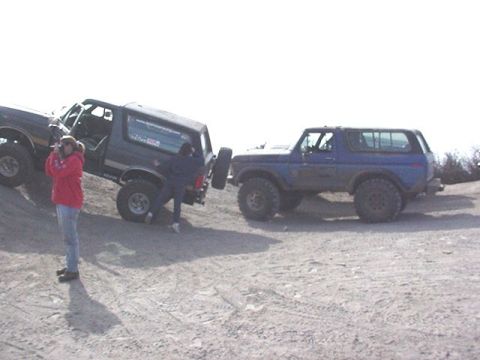 1990 Ford Bronco - Two lifted full-size SUVs parked on a dirt lot, one climbing a mound, with a person in a red jacket nearby.