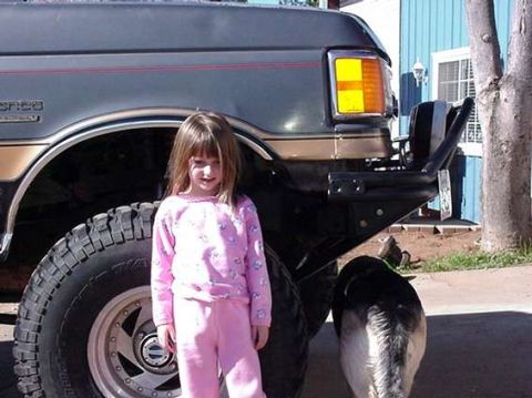 1990 Ford Bronco - young girl in pink pajamas stands beside the front fender and oversized tire of a lifted SUV outdoors.