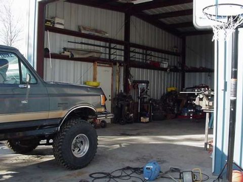 1990 Ford Bronco - lifted SUV sits inside a metal-walled workshop cluttered with tools, equipment, and a basketball hoop.